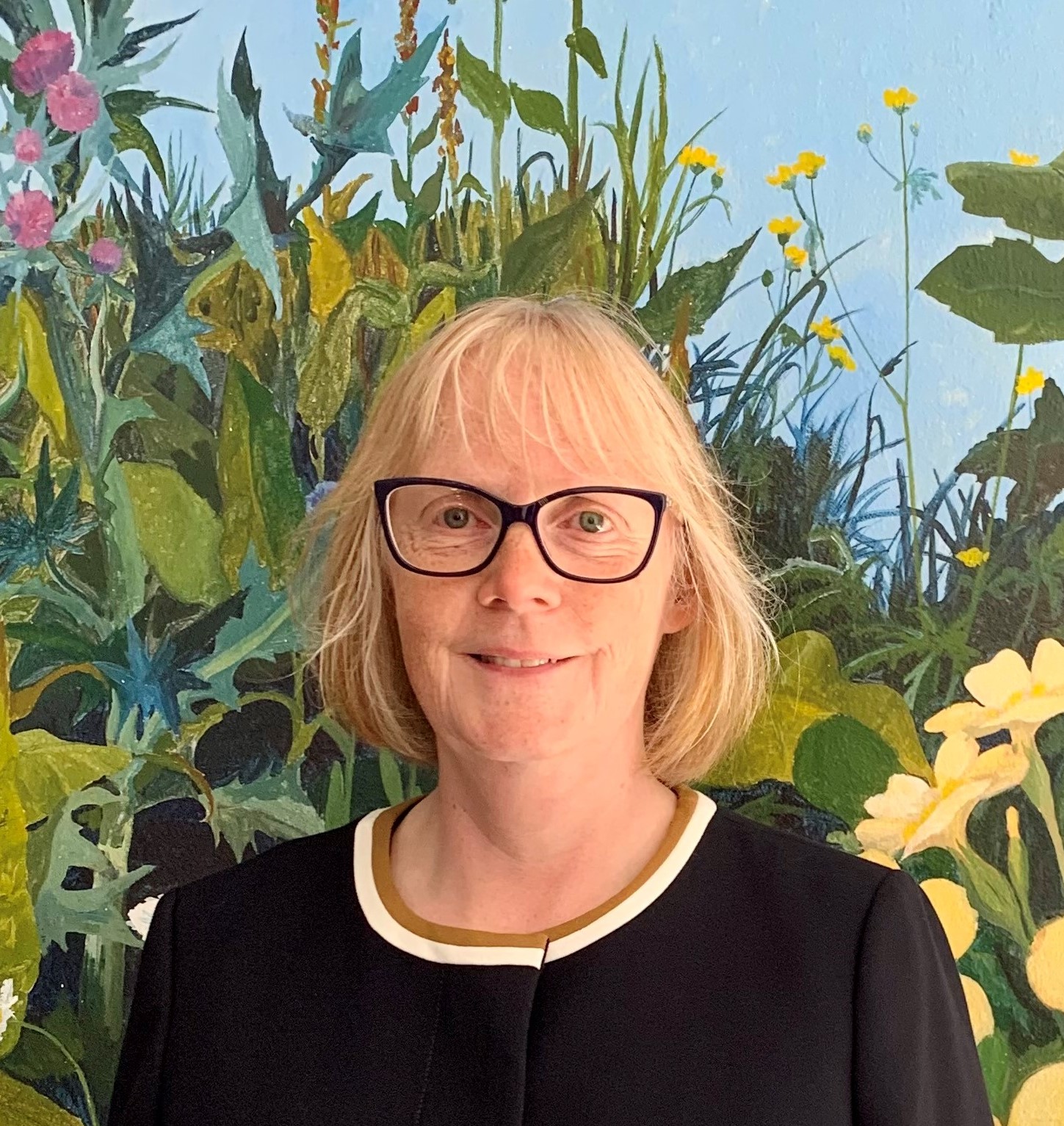 Helen Budge standing against a floral background. She is wearing dark rimmed glasses and smiling at the camera